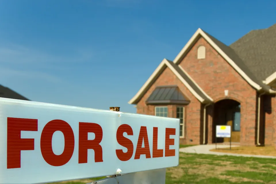 Close-up of white sign with red FOR SALE letters and blurred brick single-family home in background.